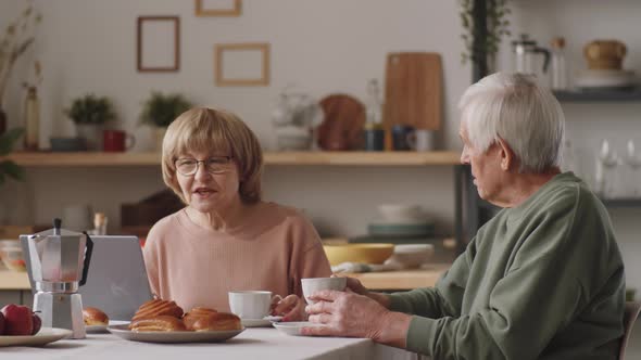 Elderly Couple Using Laptop and Speaking over Tea at Kitchen Table