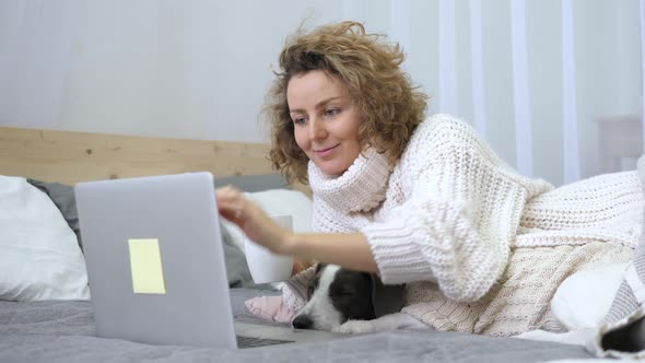 Girl In Casual Knitted Clothes Using Laptop With Dog Lying On Bed