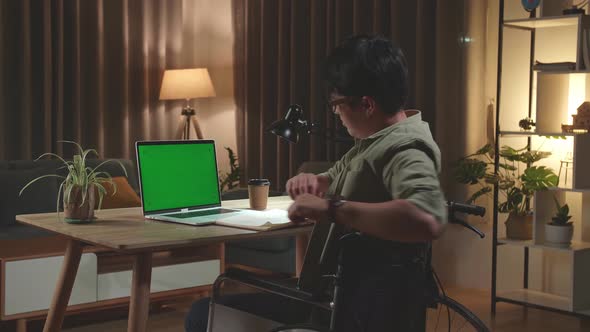 Asian Man Sitting In A Wheelchair Stretching While Working On Mock Up Green Screen Laptop Computer