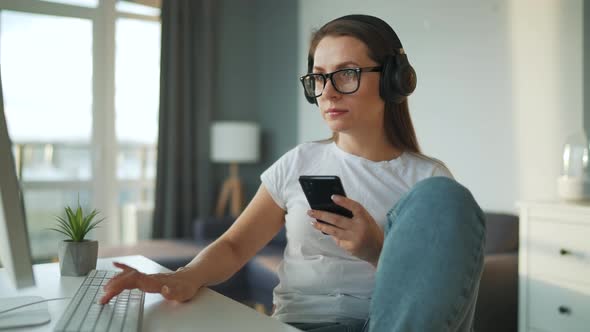 Casually Dressed Woman with Headphones Working with a Computer and Smartphone at Home in a Cozy
