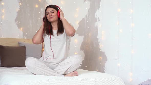 Beautiful Girl in Pajamas Listening to Music with Headphones Sitting on the Bed