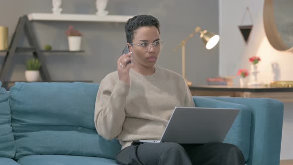 Angry African Woman with Laptop Talking on Smartphone on Sofa