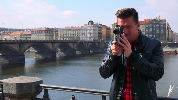 A Young Handsome Man Shoots a Video with a Camera - a River and a Quaint Townscape in the Background