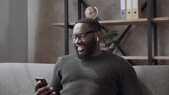 Smiling African American Man in Bluetooth Headphones Use Cellphone Rest on Comfortable Couch at Home