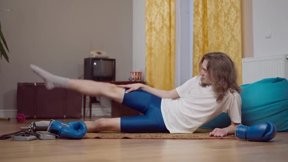Wide Shot of Skinny Concentrated 80s or 90s Man Lying on Retro Carpet Indoors Raising Leg Up