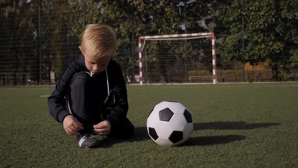 A Little Boy Soccer Player Ties His Shoelaces on a Football Field