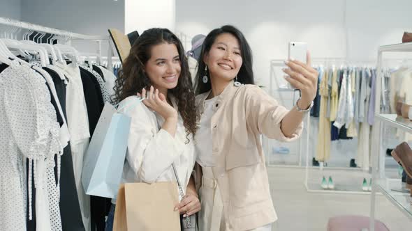 Asian and Caucasian Women Friends Taking Selfie in Clothes Shop Holding Bags Using Smartphone Camera