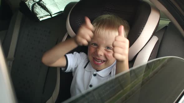 A Small Cheerful Boy Sits in a Brown Child Car Seat and Shows His Thumb Up