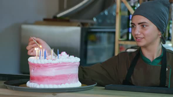 Portrait of Cute Smiling Female Baker Lights Candles on Sweet Cake To Congratulate Visitors on Their