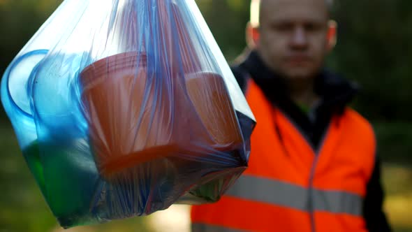 A Man in a Signal Orange Vest Holds a Package with Garbage on the Background of Nature, Forest