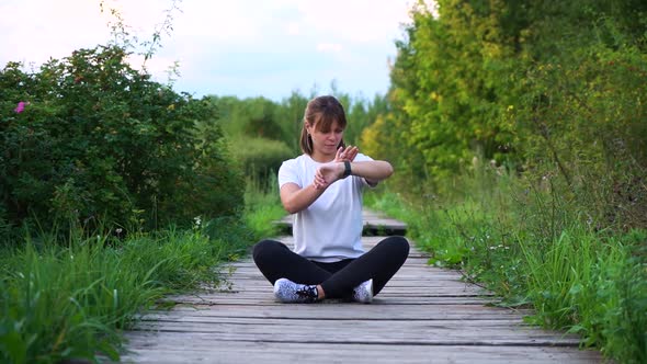 Slim Sporty Girl Sit on Wood Bridge in Lotos Pose and Look on Watch