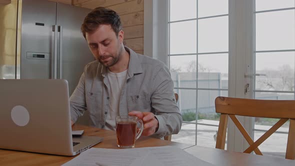 Man Drinks Tea Working at the Laptop at Home Because of Covid Restrictions