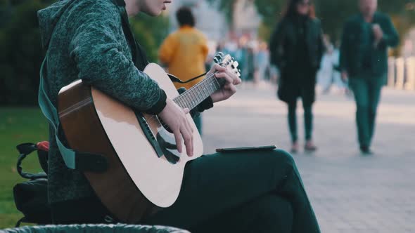 Young Street Musician Playing Acoustic Guitar in the Park. People Walk By