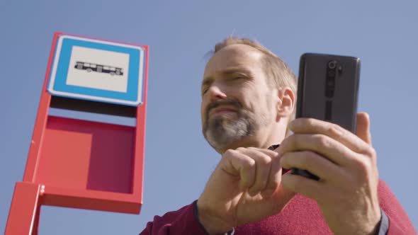 A Middleaged Handsome Caucasian Man Works on a Smartphone at a Bus Stop