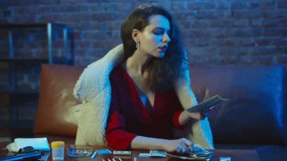 Portrait of Beautiful Young Woman Drug Dealer Sitting on Sofa Smoking Cigar and Counting Dollars