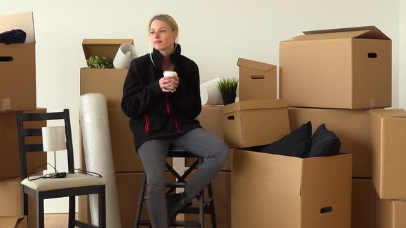 A Moving Woman Sits on a Chair in an Empty Apartment, Wearing a Jacket, and Sips a Hot Drink