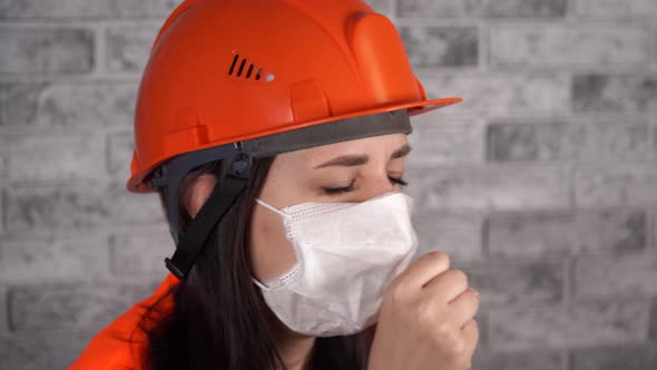 Female Construction Worker in Overalls and Medical Mask Coughing on Background of Gray Brick Wall