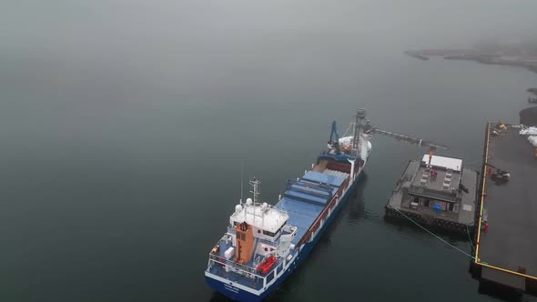 Unloading Fish From Cargo Ship On A Foggy Day In Faskrudsfjordur, East Iceland - aerial drone shot