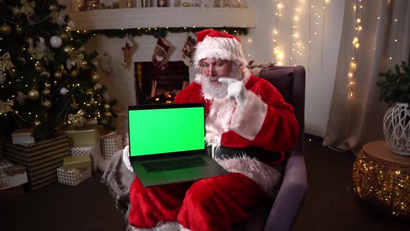 Santa Claus in Glasses Sitting in His Rocker Near Christmas Tree. Holds a Laptop with Green Screen