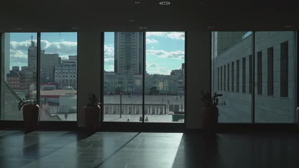 Man Goes Down a Long Corridor Room with High Windows and Flowerpots