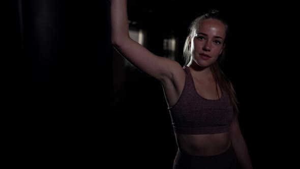 Confident Athletic Woman Standing at Punching Bag in Darkness Looking at Camera