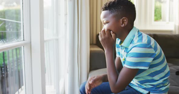 Stressed african american boy looking out of the window while sitting on the couch at home