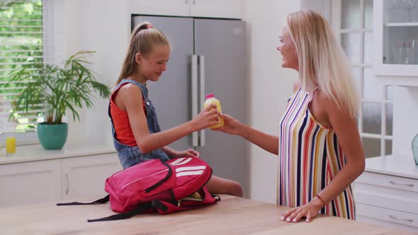 Smiling caucasian mother in kitchen with daughter preparing school bag, packing juice and talking