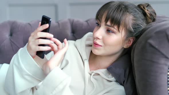 Portrait of Young Attractive Woman Enthusiastically Chatting Using Smartphone Lying on Couch