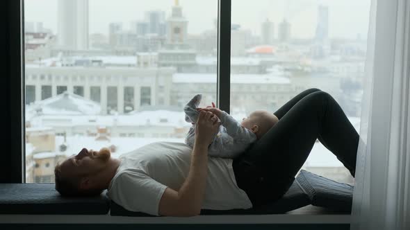 baby lies with dad at a large window overlooking the city