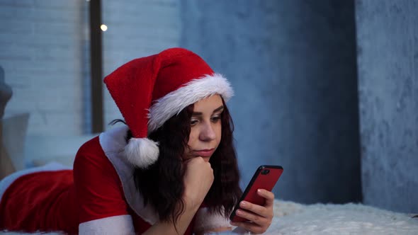 Young Woman in Santa Claus Costume Browses Smartphone, Lying on Bed. Charming Female Resting with