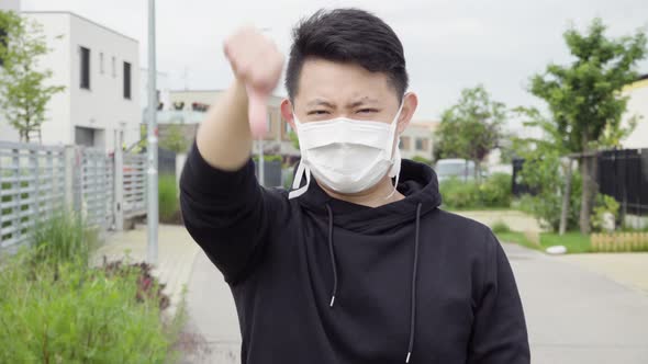 A Young Asian Man in a Face Mask Shows a Thumb Down To the Camera and Shakes His Head