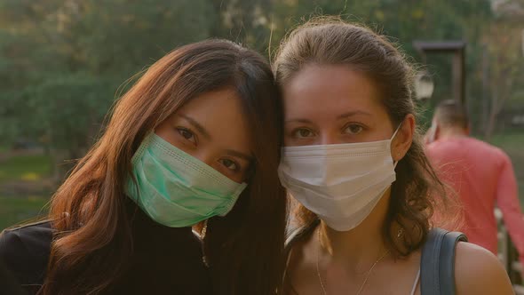 Portrait of Two Young Women in Love Wearing Protective Face Medical Mask Standing in Park at Sunset