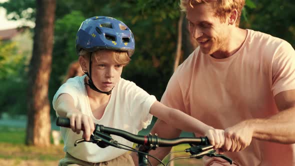 A happy father is helping his son to ride the bicycle while walking