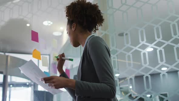 Mixed race businesswoman writing with green marker on memo note on transparent board in office