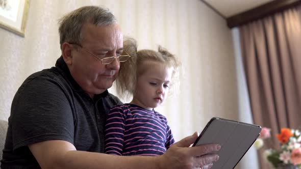 Granddaughter and Grandfather Are Sitting with a Tablet. The Girl Frowns at the Tablet. Sitting on