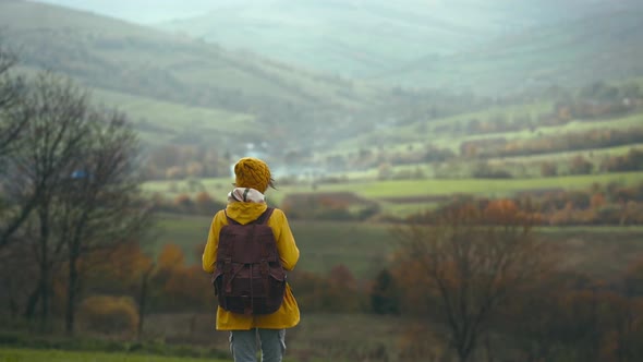 Woman Hiker with Backpack Walking By Green Meadow Among Hills Girl Wearing Yellow Jacket and Hat