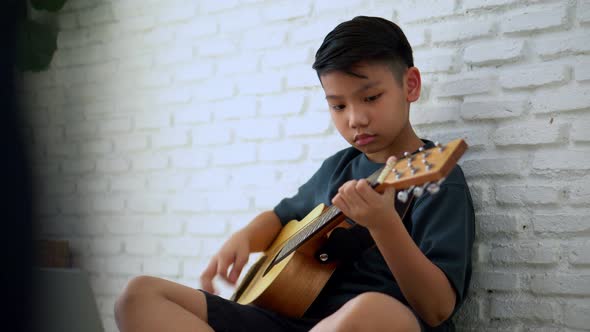 Preteen Asian boy playing acoustic guitar dressed sitting on the cozy sofa at the home living room a alt
