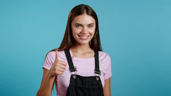 Cute Woman Showing Thumb Up Sign Over Blue Background. Positive Young Girl Smiles To Camera. Winner