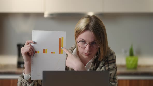 A Young Woman with Glasses at Home Shows a Graph to a Laptop Camera While Sitting at Home in the