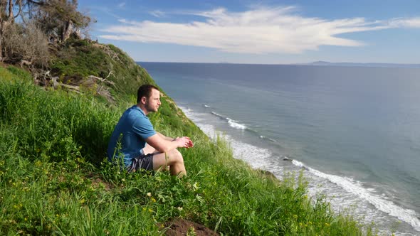 A young man traveler sitting down and enjoying nature in a grassy field on the edge of a beach cliff