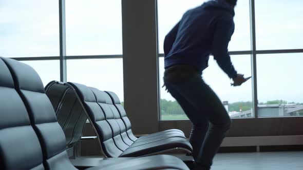 Man Comes in an International Airport Lobby and Sits Down on Bench with Smartphone in His Hand