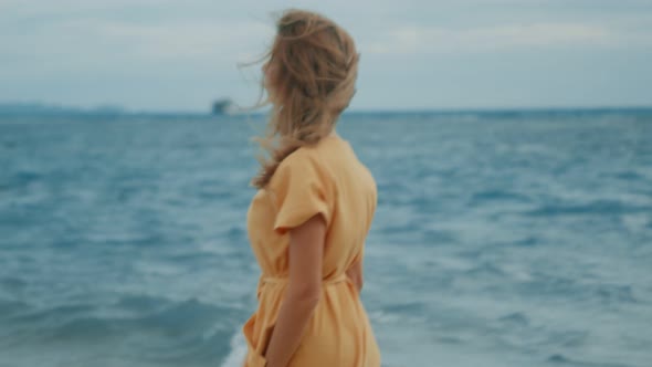 A Slim Blonde in a Yellow Dress Walks Along the Beach and Looks Around