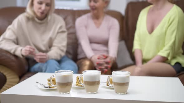 Three Cups of Cappuccino and Cakes on Table with Blurred Unrecognizable Adult Friends Talking