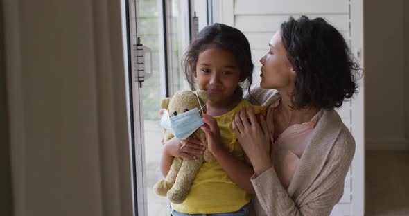 Hispanic mother and daughter embracing in window holding teddy bear with face mask