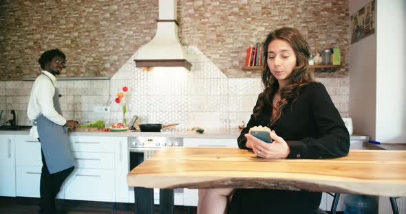 Young Black Man Cooking and Talk with Latin Wife Sitting at Table Using Smart Phone in Kitchen