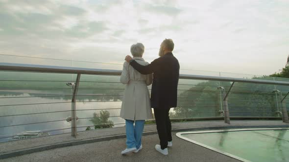 Old Man Hugging Mature Wife Standing on Bridge Admiring Park and River