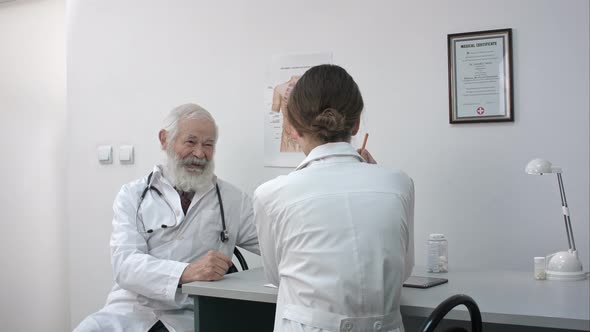 A Senior Male Doctor at a Desk in an Office Smiling and Talking To Female Doctor