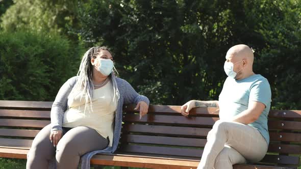 Pandemic Concept. Two Happy Friends in Medical Mask Keep Social Distance Sitting and Talking Outside