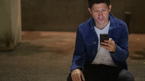 Hispanic Man Thinking and Using Phone While Sitting on the Street in the City at Night