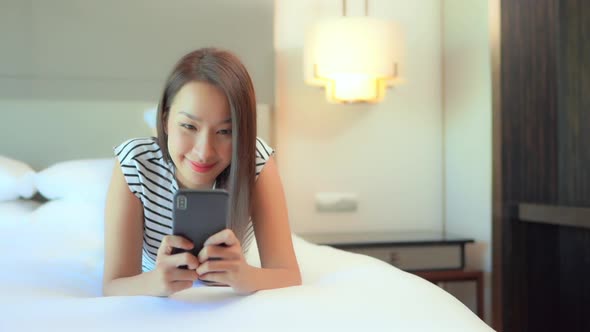 Texting on Smartohone in Bed. A Young Happy Asian Woman in Bedroom Chatting With Mobile Phone, Slow